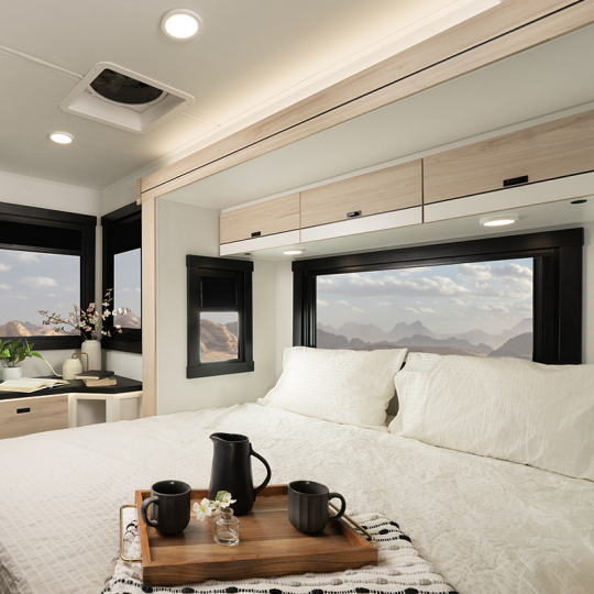 A cozy bedroom in a modern RV, featuring a plush bed with white bedding, a wooden tray with a coffee pot and two mugs, and large windows showcasing a mountain view. Decorative elements include a flower vase and a small desk area.