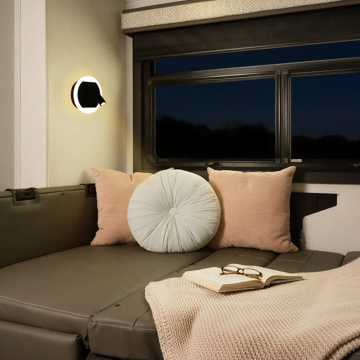 Cozy living space featuring a gray sofa with plush pink and round decorative pillows, a warm knitted blanket, an open book, and a modern wall lamp illuminating the area beside a large window at dusk.
