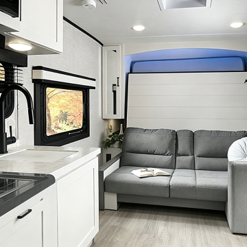 Modern camper interior featuring a gray sofa, kitchen area with white cabinetry, and a scenic view through the window. A book rests on the sofa, enhancing the cozy atmosphere.