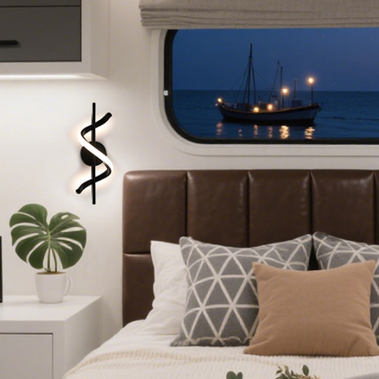A cozy bedroom with a brown leather headboard, light-colored decorative pillows, and a potted plant, featuring a view of a boat with lights at dusk through a window, along with a modern wall lamp shaped like a dollar sign.