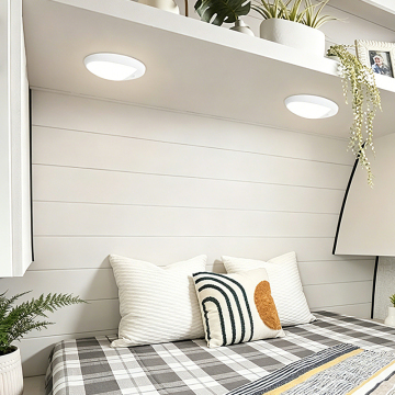 A cozy bedroom corner featuring a modern bed with a plaid patterned blanket, decorative pillows, a white shelf with plants and a framed photo, and soft ceiling lights illuminating the space.
