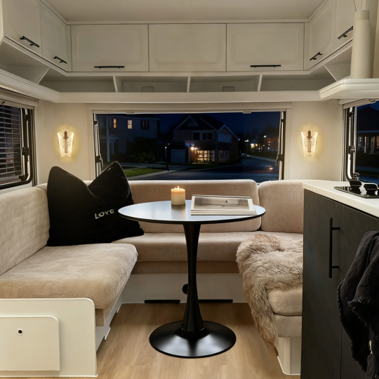 A cozy RV interior featuring a circular black table, plush beige seating, a candle on the table, and a stylish throw blanket, illuminated by soft lighting in the evening.