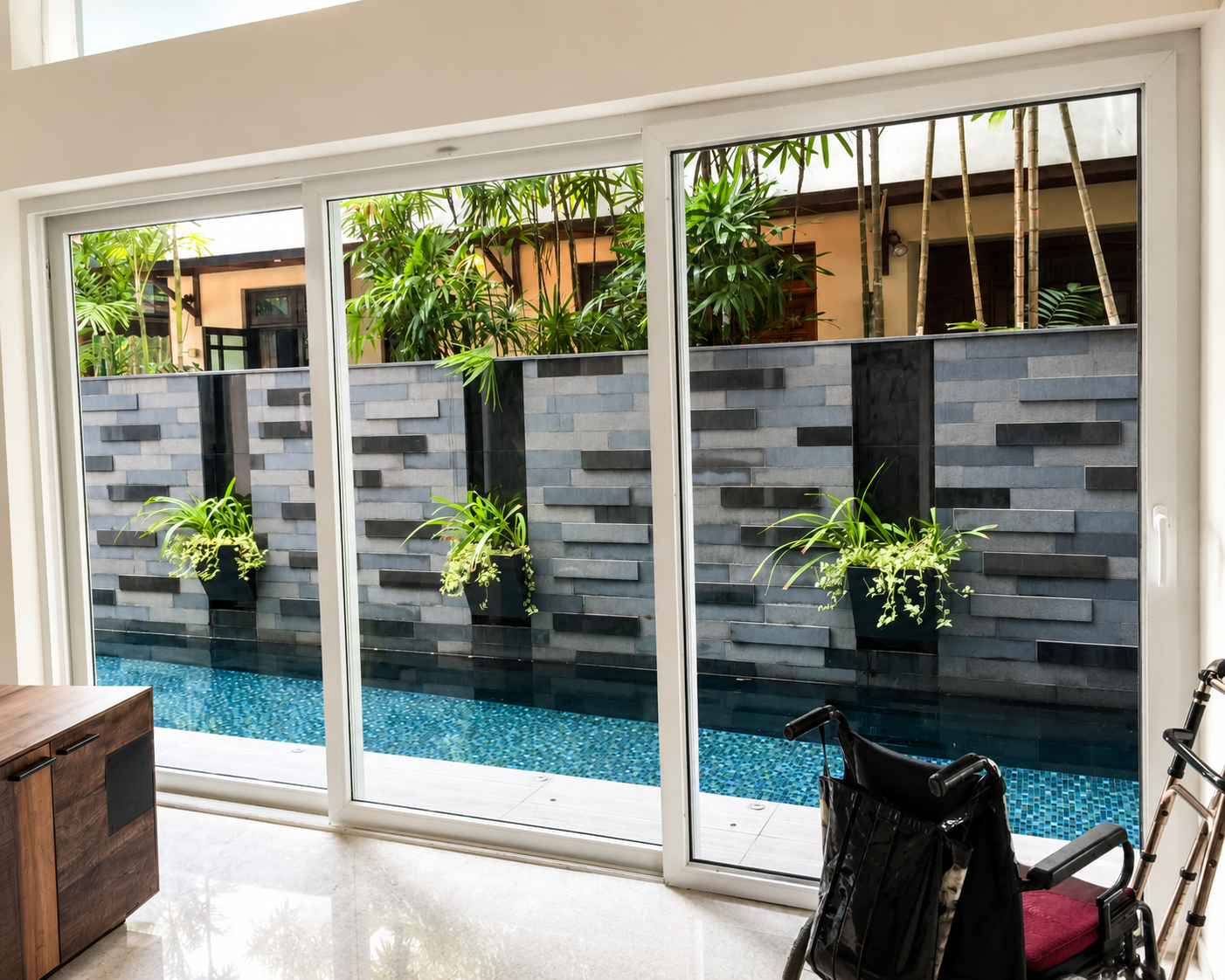 A modern living space featuring large glass doors that open to a pool area, with a sleek stone wall adorned with green plants. A wheelchair is positioned nearby, creating an inviting and accessible atmosphere.