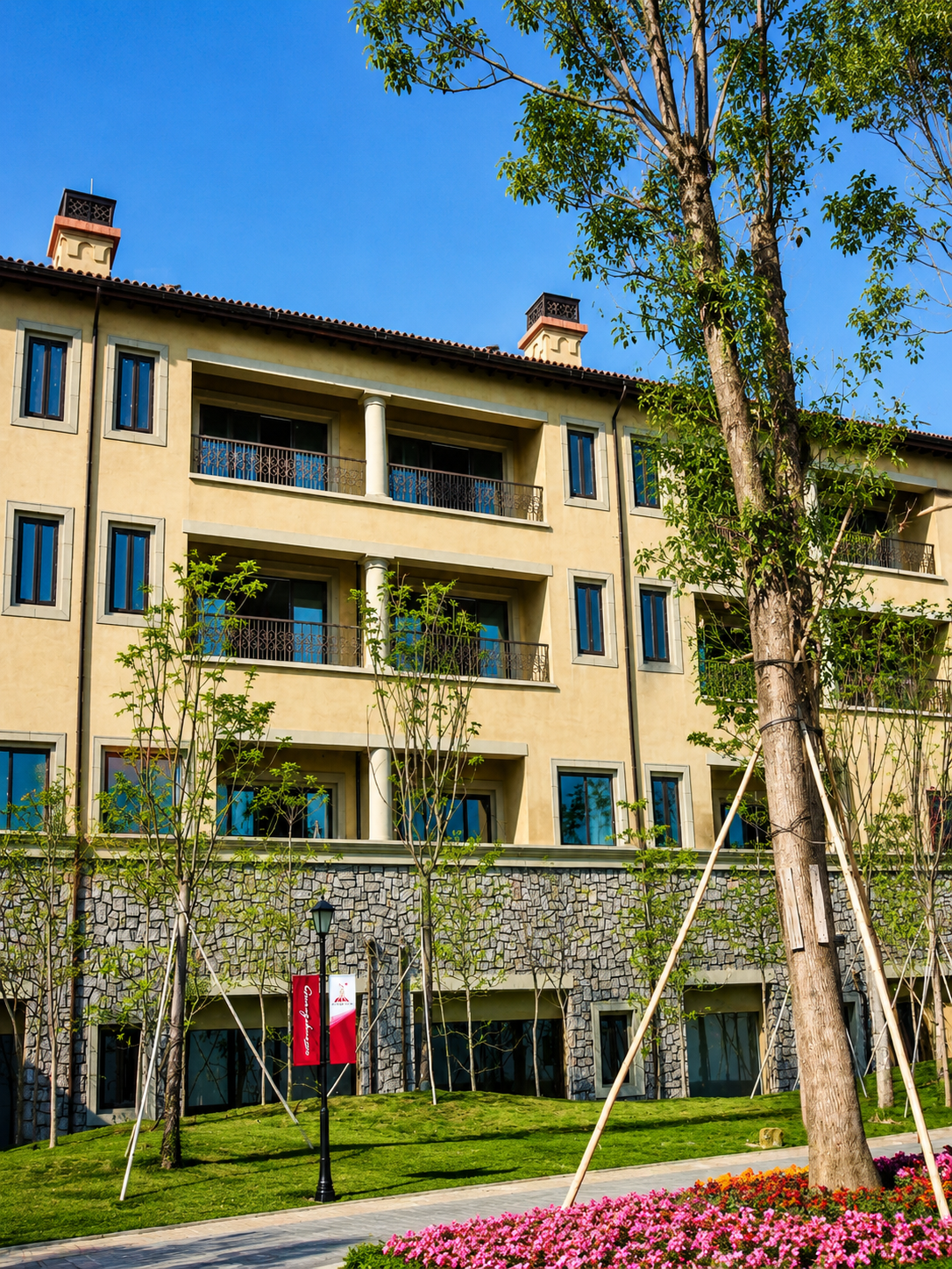 A modern multi-story building with balconies surrounded by lush greenery and colorful flower beds under a clear blue sky.