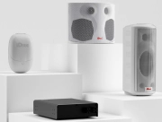 White speakers and a black audio amplifier from iDste displayed on white pedestals in a minimalist setting.