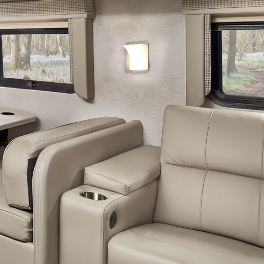 A cozy interior of a vehicle featuring light beige leather seating, a cup holder, and soft wall lighting, with a view of a forest visible through the window.