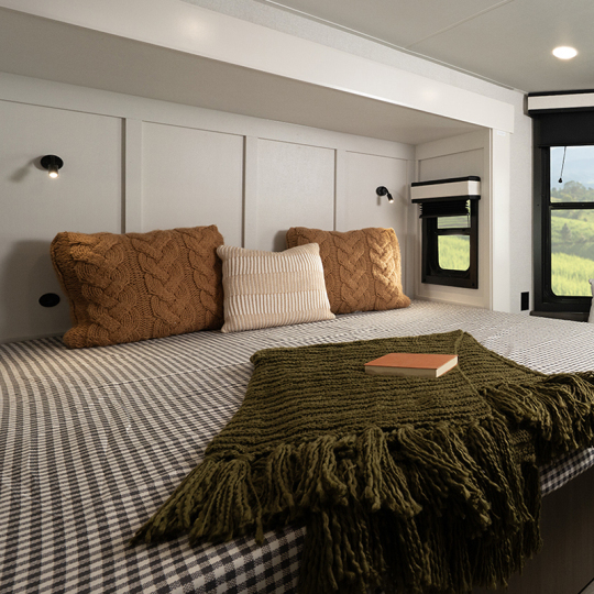Cozy interior of a minimalist bedroom featuring a checkered bedspread, two knitted throw pillows in brown and cream, an olive green blanket, and a wooden book on the bed. Natural light streams in through a nearby window.