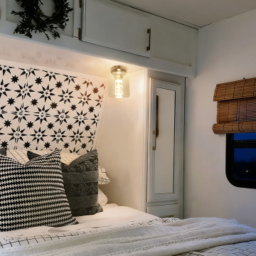 A cozy bedroom featuring a geometric patterned headboard, decorative pillows in black and white, wall-mounted light fixture, and natural bamboo window blinds.
