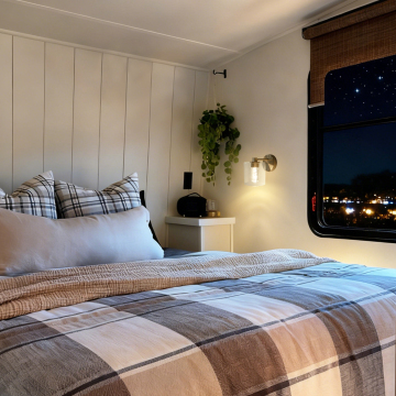 A cozy bedroom featuring a comfortable bed with plaid bedding, decorative pillows, a potted plant hanging on the wall, and a charming wall light, illuminated at night with a view of city lights through the window.