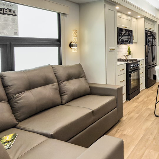 Modern living room featuring a brown leather sofa, stylish wall light, and a kitchen area with dark appliances, white cabinetry, and stylish decor accents.