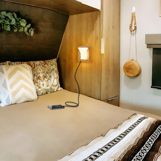 Warm white RV wall reading light beside a bed, illuminating a cozy camper bedroom with a smartphone charging below